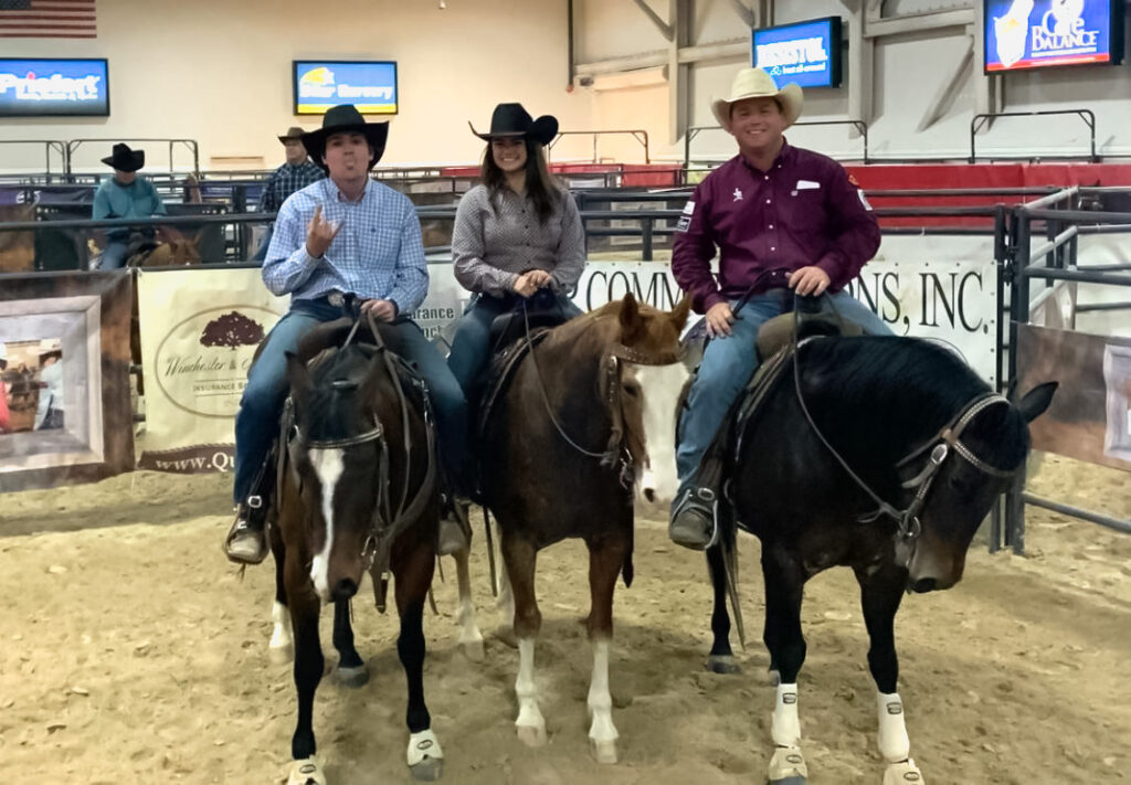 Three riders in cowboy hats sit mounted on their horses side by side in an indoor arena, smiling for the camera
