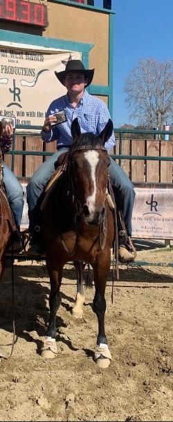 Cowboy in a black hat and plaid shirt holds up a trophy buckle while mounted on a bay horse in an arena