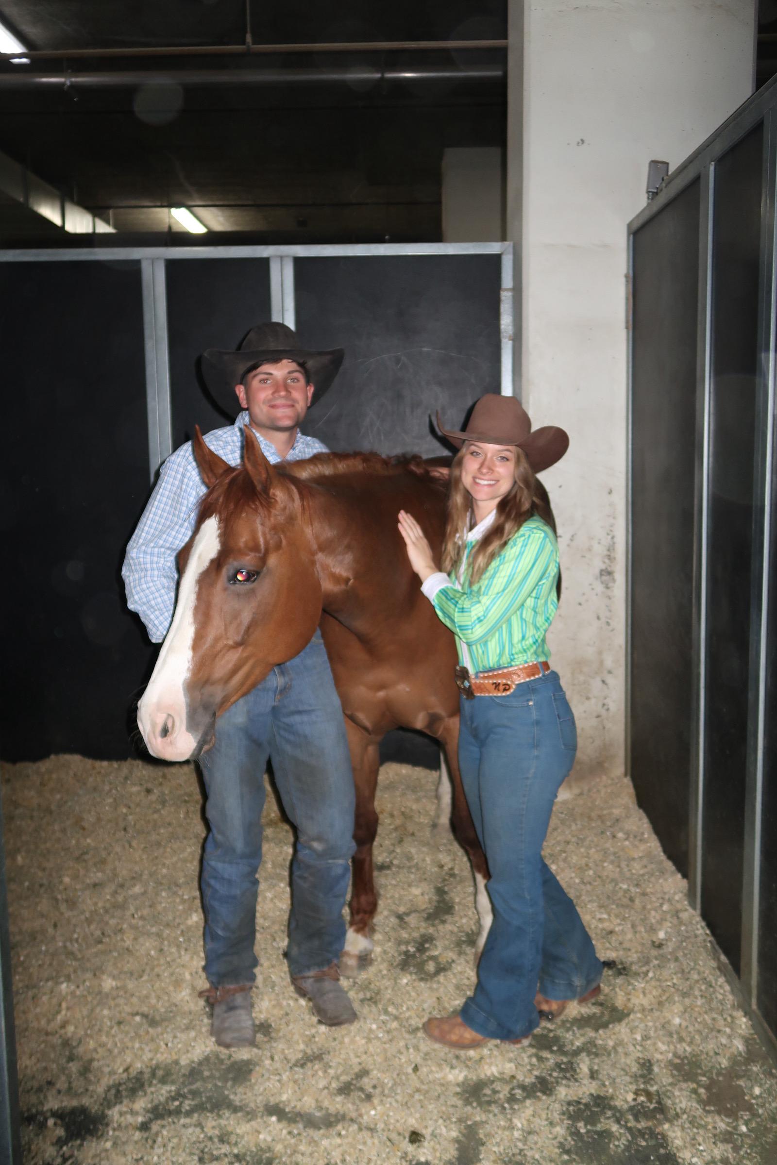 Two smiling young people in cowboy hats stand beside a chestnut horse with a blaze in a barn alleyway