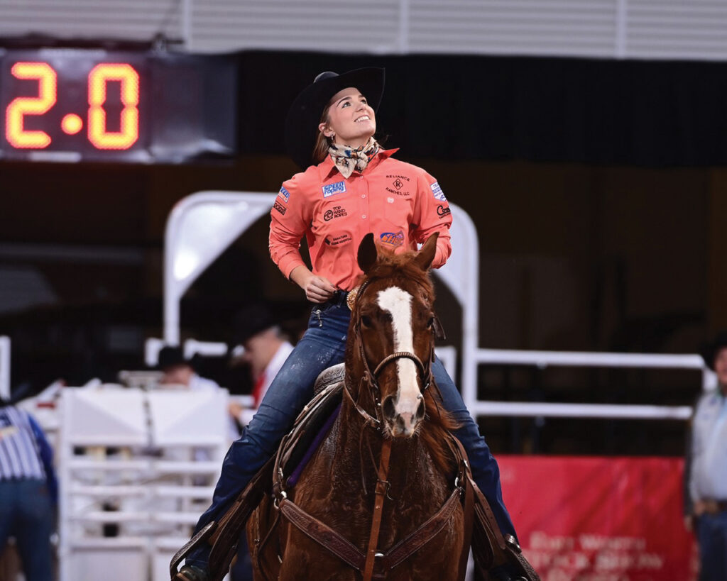 Smiling breakaway roper in a coral shirt and black cowboy hat rides her sorrel horse past a scoreboard reading 2.0