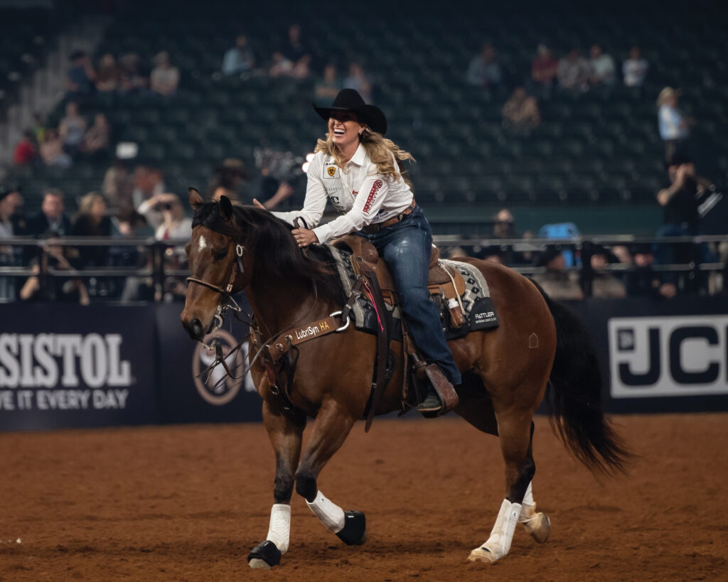 Smiling cowgirl in a black hat and white shirt rides her bay horse across the arena after a rodeo run