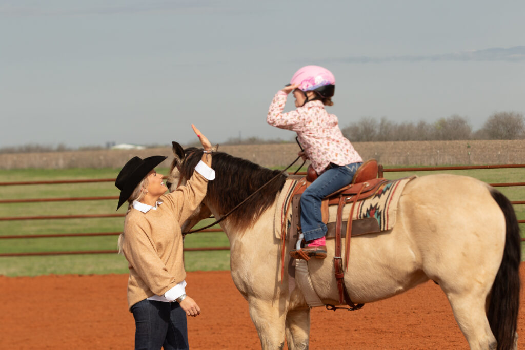 Young rider in pink helmet adjusts saddle while handler in tan shirt and black cowboy hat holds calm palomino horse in red dirt arena