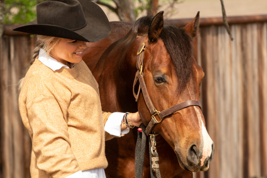 Cowgirl in tan shirt and black cowboy hat holds halter of brown horse with white blaze against metal barn backdrop