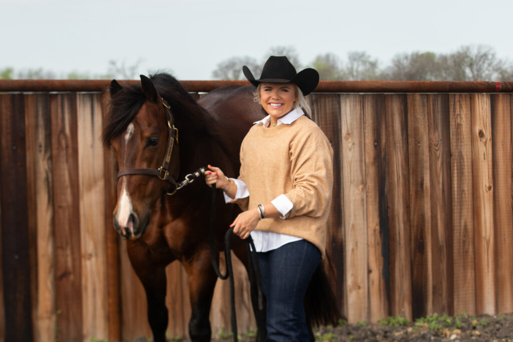 Woman in cowboy hat holding lead rope of bay horse with white blaze against wooden fence
