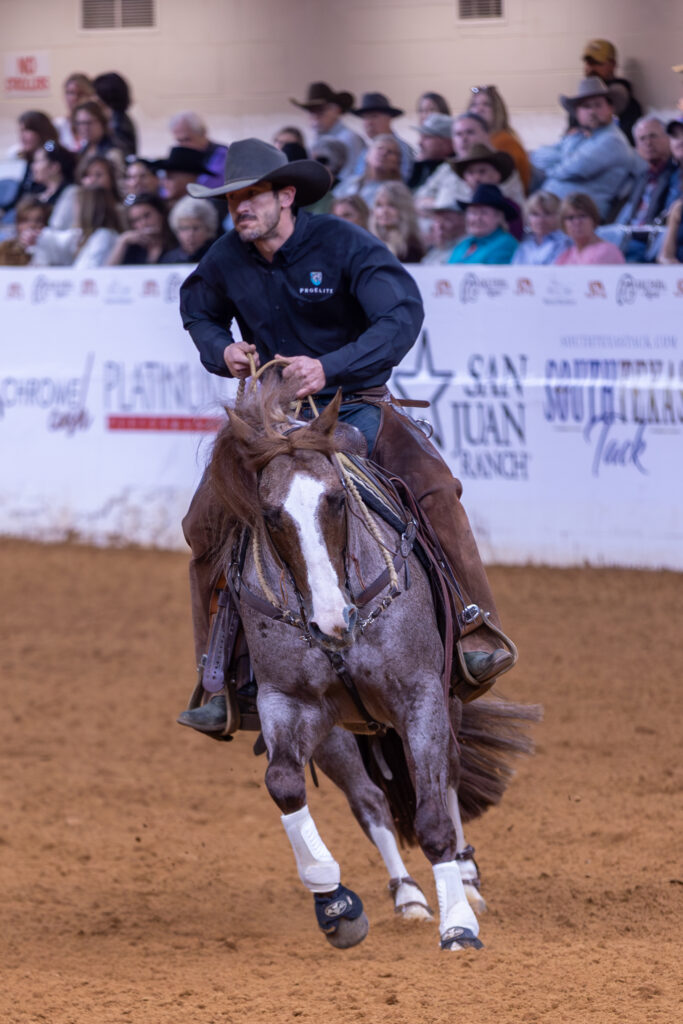 Cowboy in navy shirt riding a bay horse with white blaze across an arena dirt floor during a rodeo event with spectators in bleachers behind