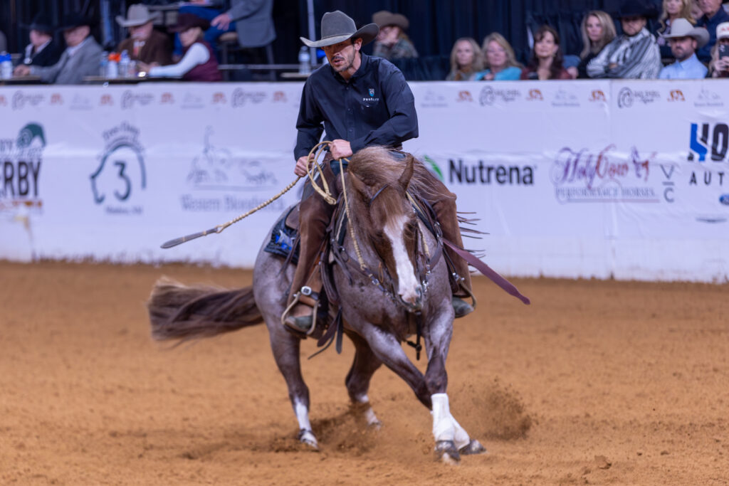 Rider in dark shirt on brown horse with white markings performing reining maneuver in arena with sponsor banners visible
