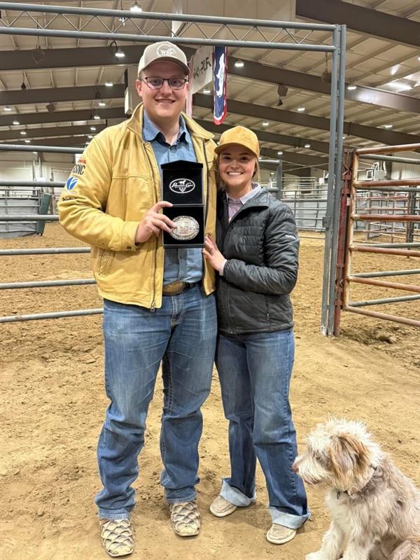 Two people hold a Toyota belt buckle award in an indoor arena with a dog at their feet