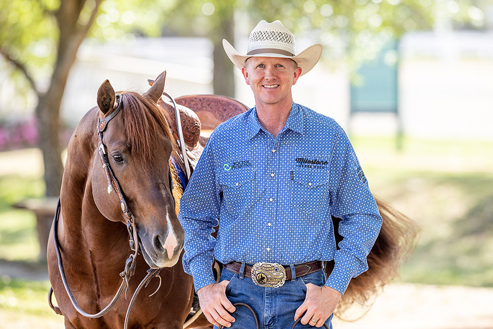 Cowboy in blue shirt and white hat posing with brown horse at outdoor ranch setting