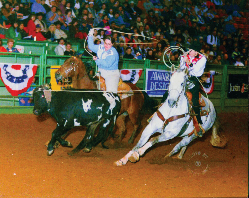 Two team ropers swing their loops as they chase a black-and-white steer across the arena in front of a packed crowd