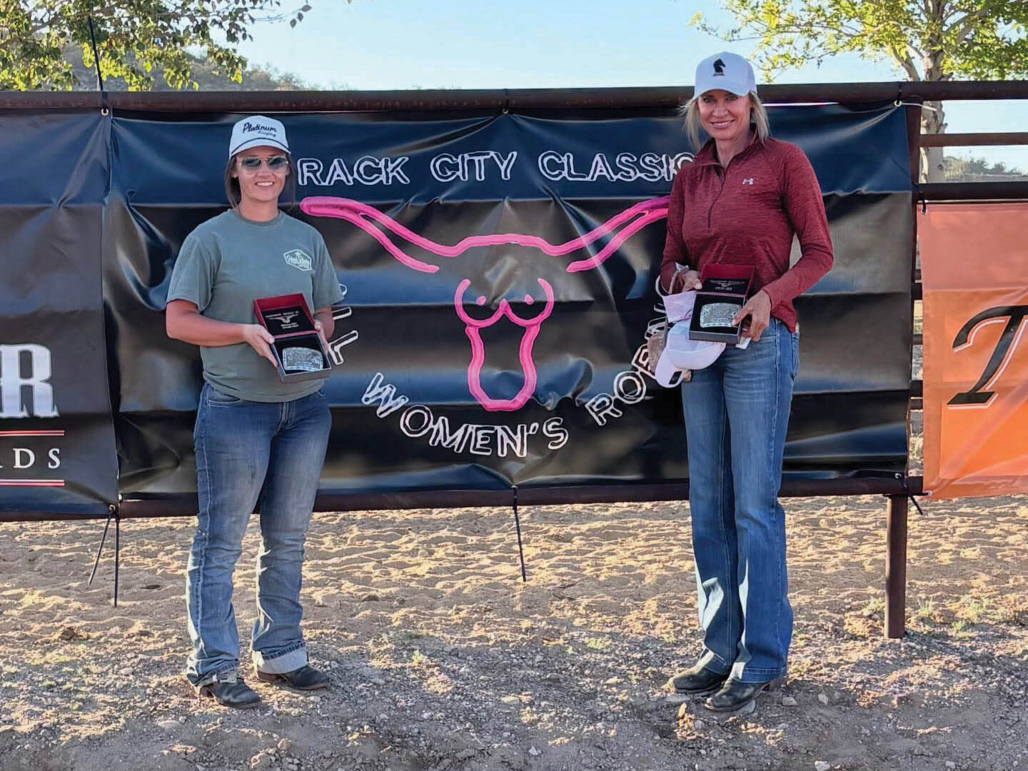 Two women in ball caps hold open buckle boxes in front of a Frack City Classic women's rodeo banner