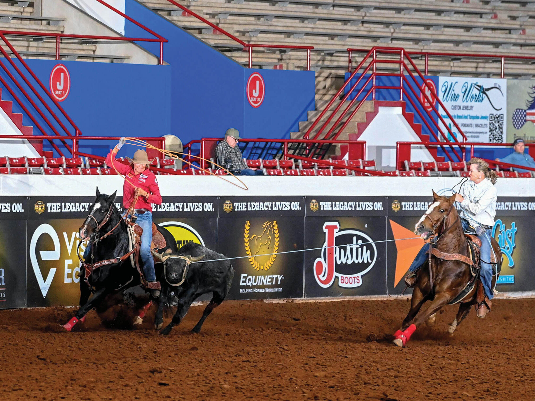Two women team ropers in an indoor arena, the header on a sorrel holding the steer while the heeler on a black horse swings her loop
