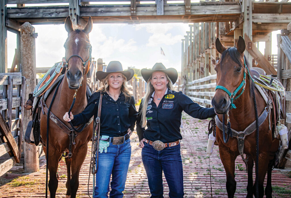 Two smiling women in cowboy hats and black shirts stand between their saddled horses in a brick-floored rodeo alleyway