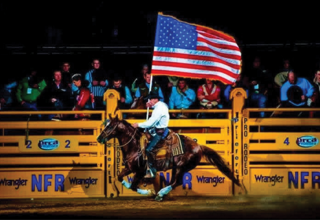 Cowboy in white shirt gallops a bay horse carrying the American flag past Wrangler NFR arena banners