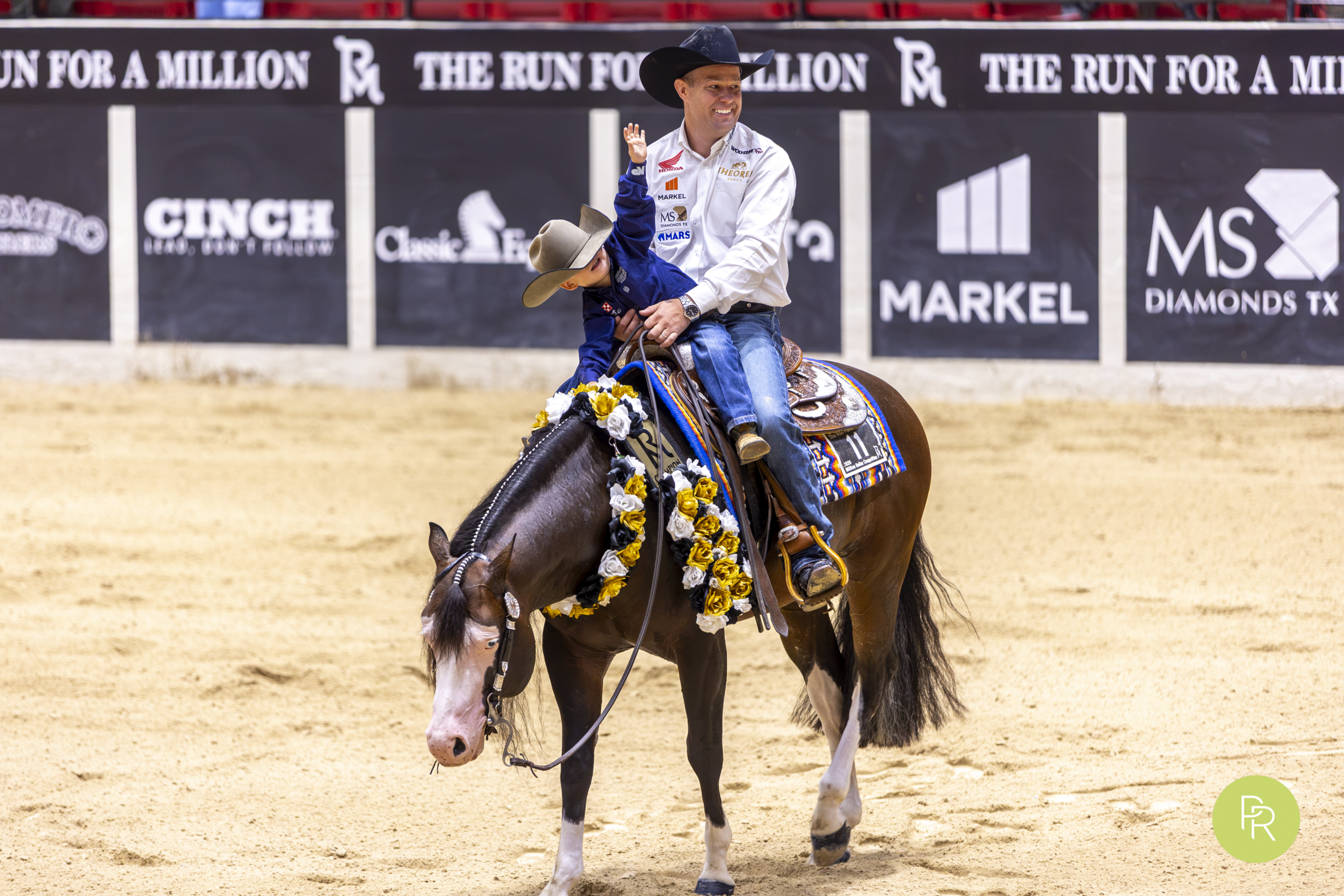 Rider in blue shirt on black horse with yellow saddle blanket in arena with sponsor signage during rodeo event