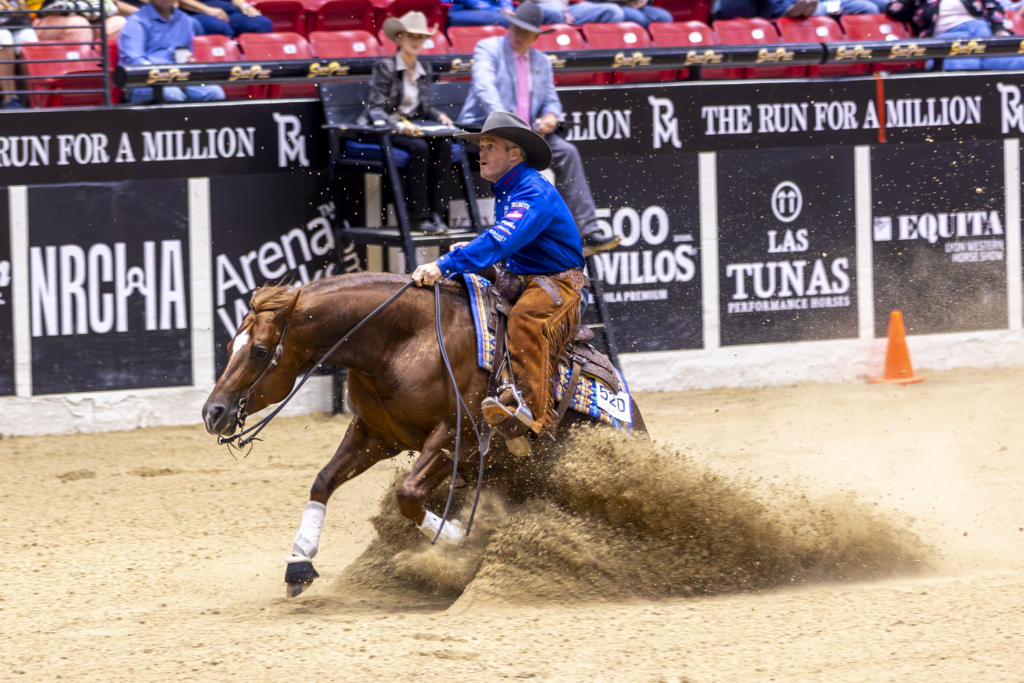 Barrel racer in blue shirt leans into turn around barrel, horse kicking up sand in indoor arena