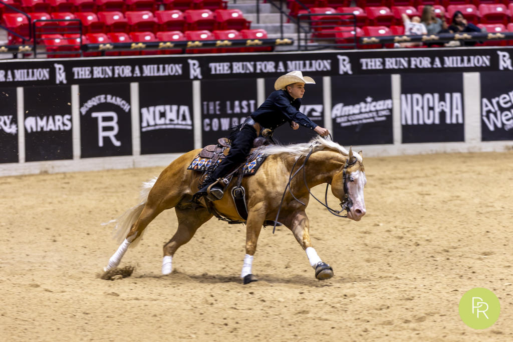 Rider in dark shirt on tan horse turning left across arena dirt at indoor rodeo venue with red seats and sponsor banners in background