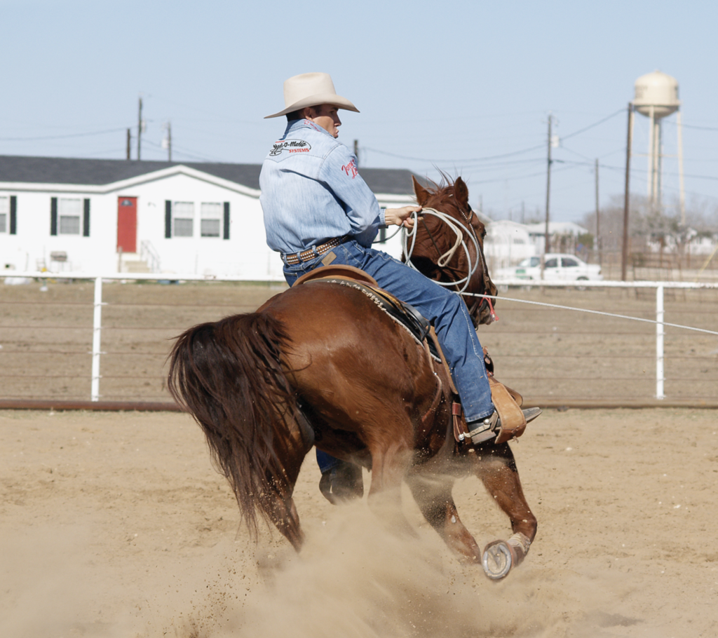 Cowboy in blue shirt and tan hat gallops brown horse through dust in arena with white fencing and ranch buildings in background