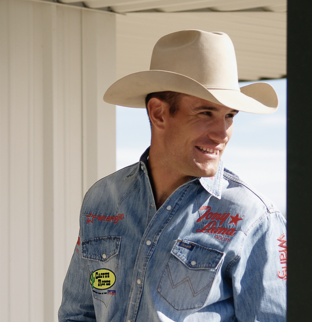 Professional rodeo competitor in denim shirt with sponsor logos wearing cream cowboy hat, smiling indoors