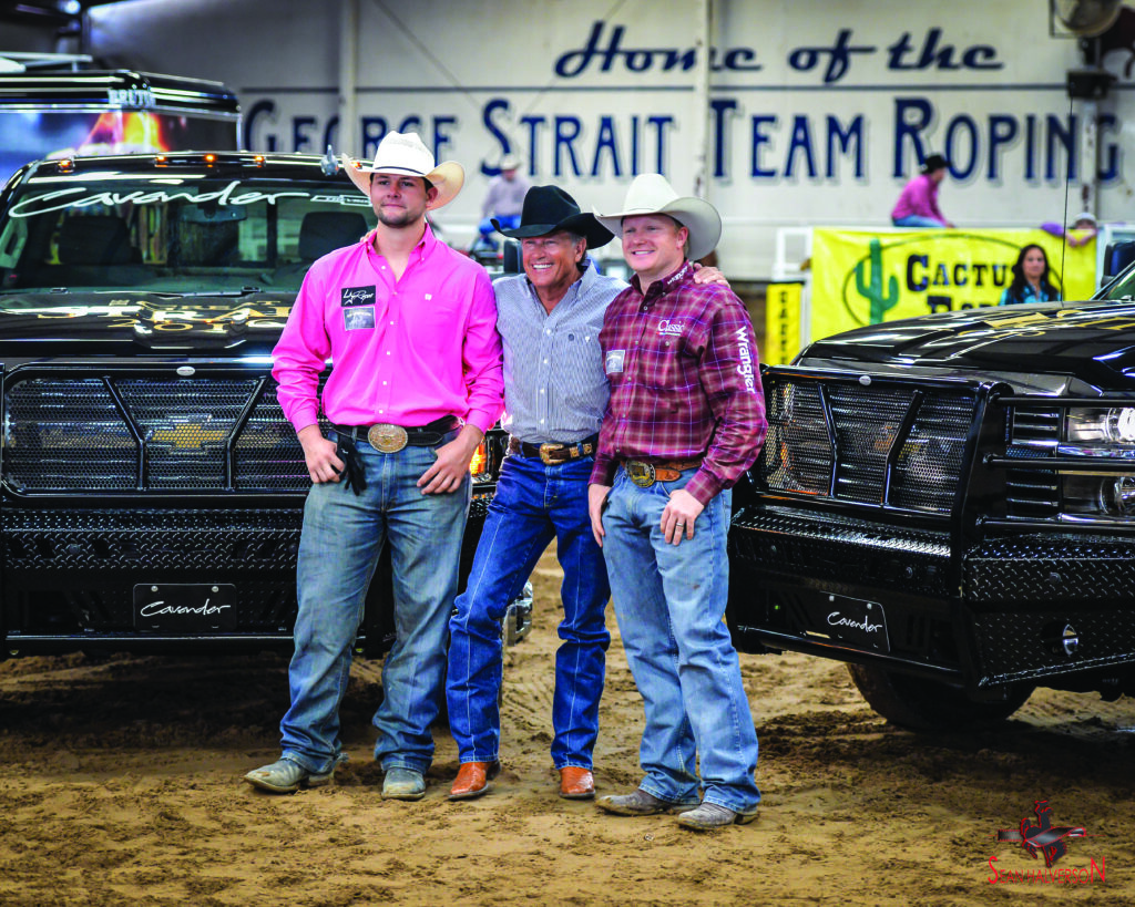 Three men in cowboy hats stand arm in arm between two black Cavender's pickup trucks beneath a George Strait Team Roping banner