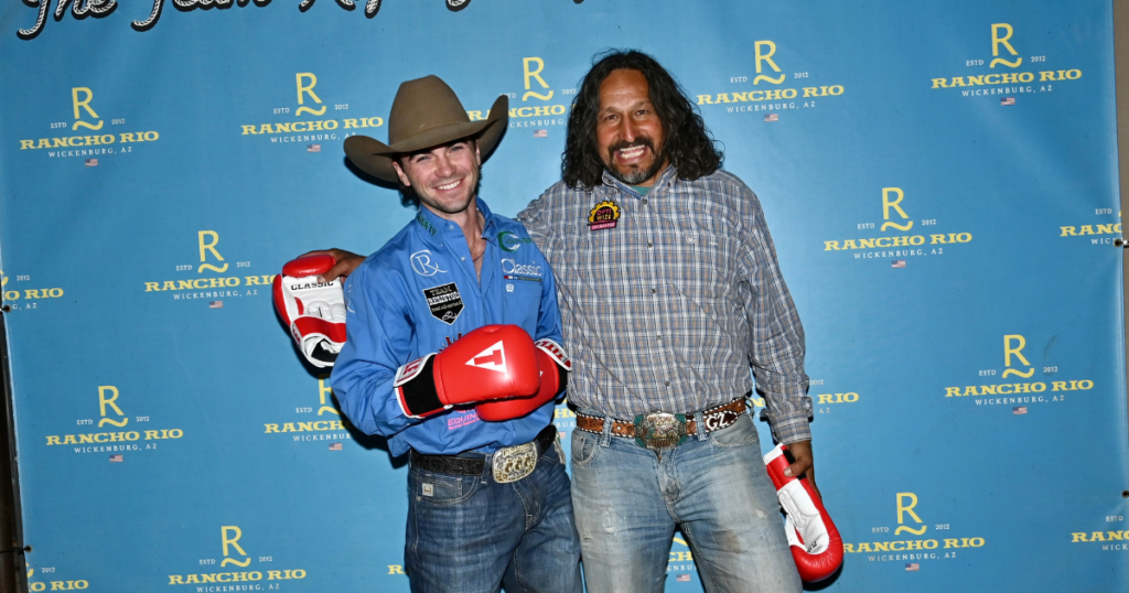 Two men pose at a Rancho Rio event, one wearing a blue jacket and cowboy hat holding red boxing gloves, the other in a plaid shirt