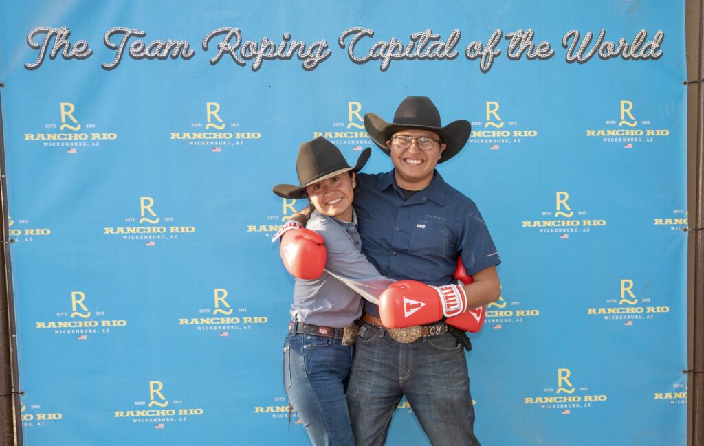 Two people in cowboy hats and red boxing gloves pose together at Rancho Rio event backdrop in Wicksburg, Arizona