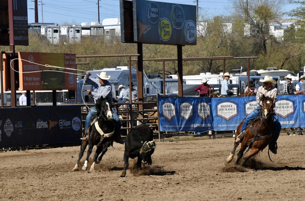 Team ropers on horseback work a steer in the arena at a rodeo sponsored by Shadle Insurance, with spectators and trailers visible in the background