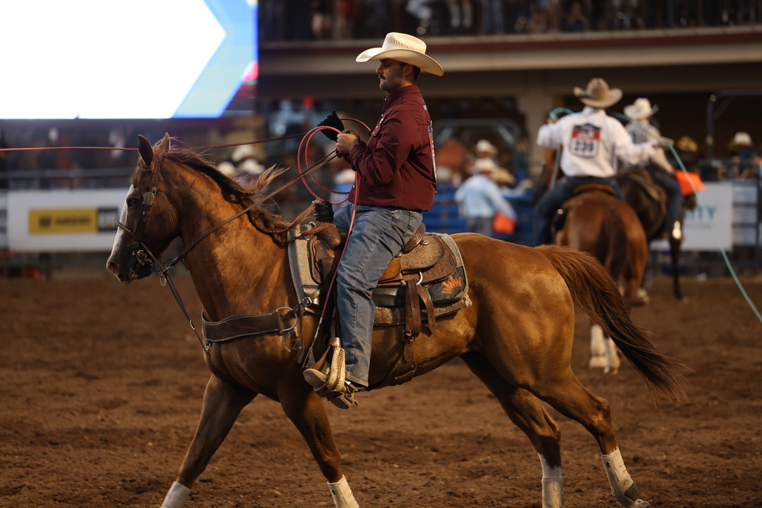 Cowboy in a maroon shirt and straw hat coils his rope while riding a sorrel horse across the arena dirt