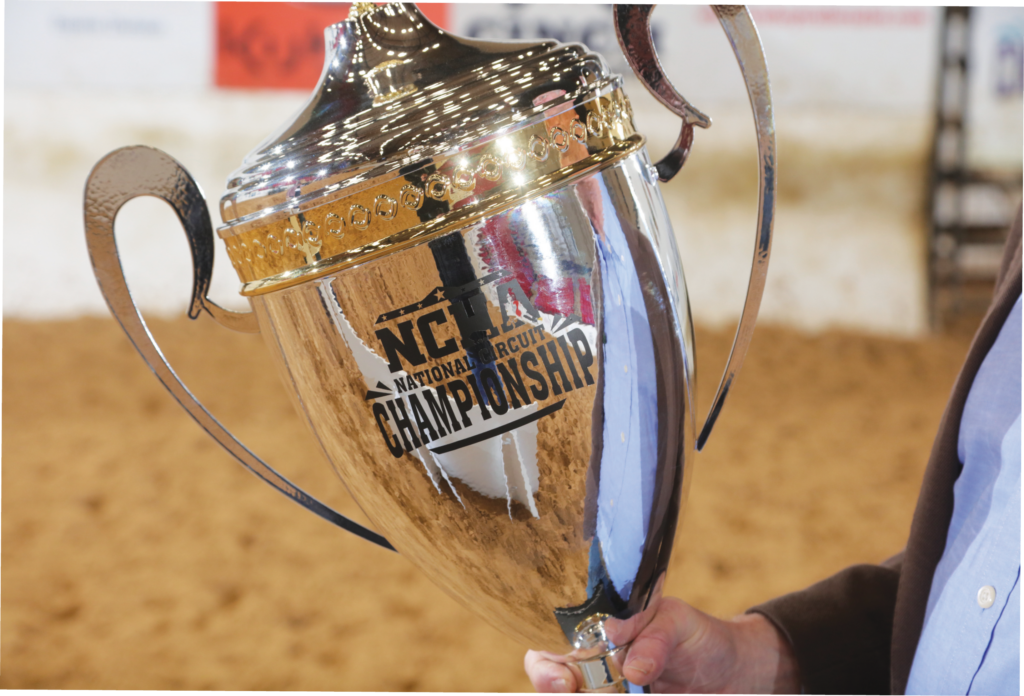 Hand holding a large silver and gold NCHA National Circuit Championship trophy in a dirt arena