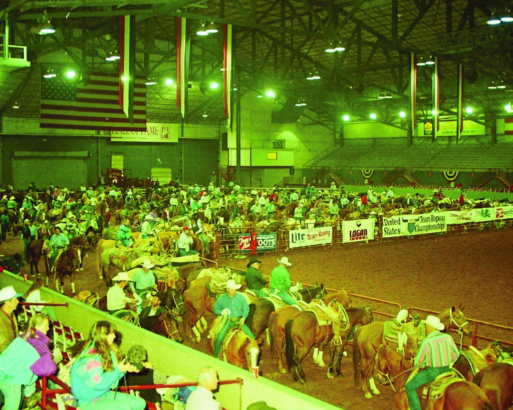 Cowboys and cowgirls on horseback compete in team roping at an indoor rodeo arena filled with spectators under green lighting