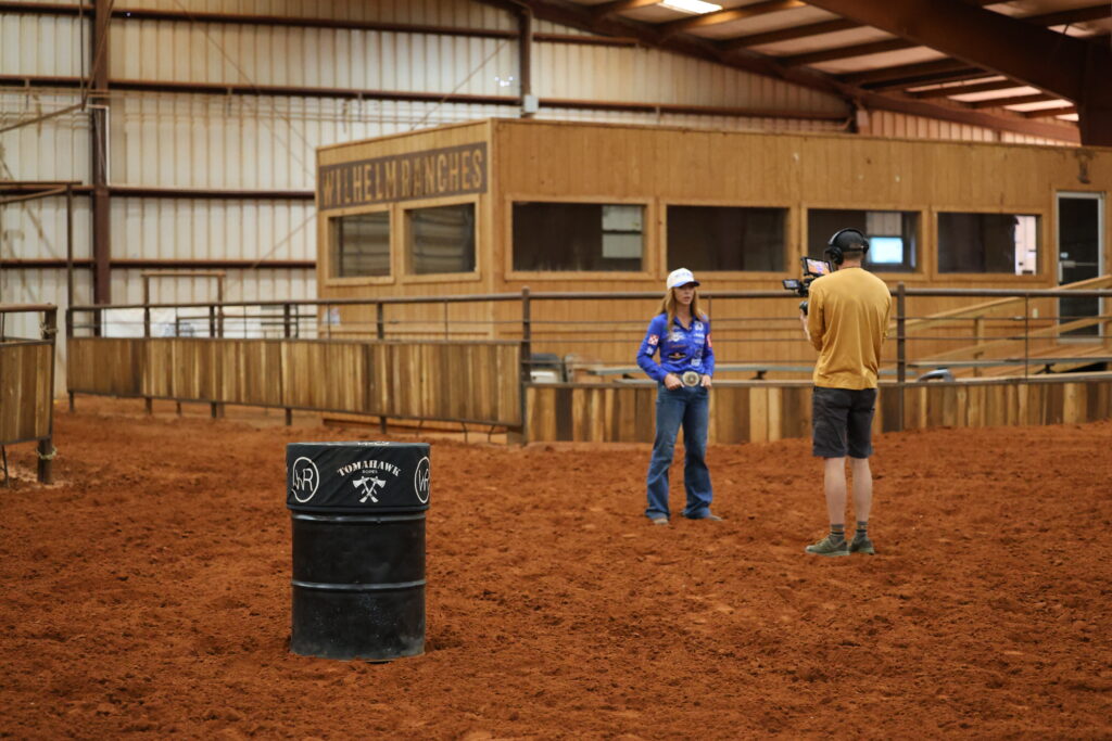 Barrel racer in a blue jersey holds her trophy buckle while being filmed by a videographer in an indoor arena