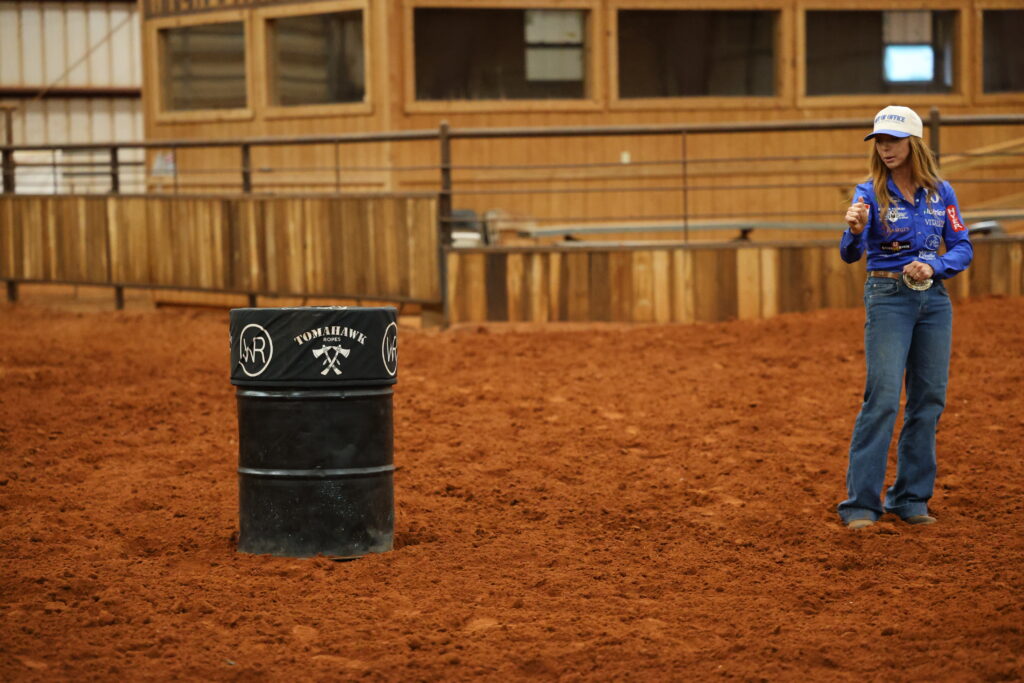 Woman in a blue sponsor shirt and ball cap demonstrates a roping grip beside a Tomahawk Ropes barrel in a dirt arena