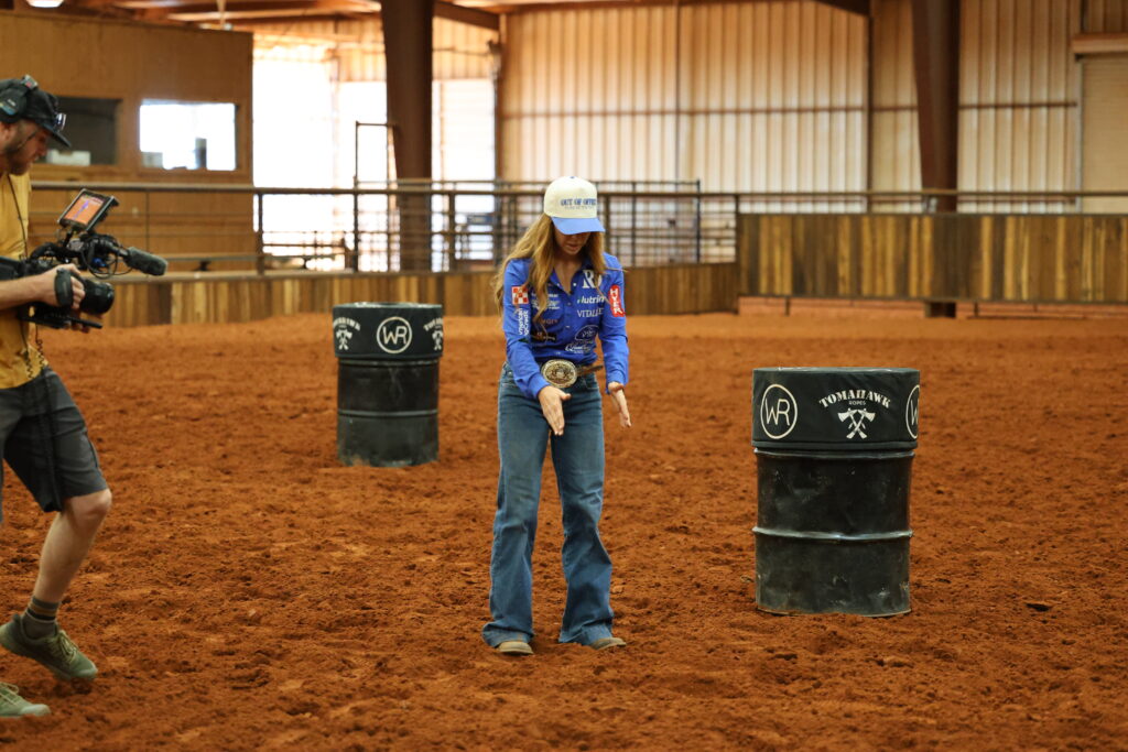 Barrel racer in a blue shirt and ball cap walks a pattern between WR-branded barrels in a covered arena as a videographer films