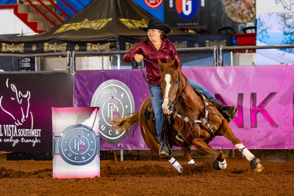 Barrel racer in a maroon shirt and black cowboy hat turns her sorrel horse around a Pink Buckle barrel in an indoor arena