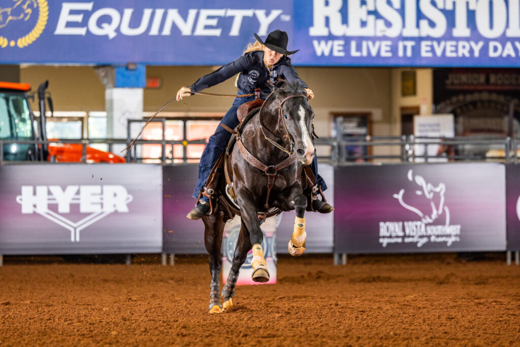 Breakaway roper in a black hat leans forward swinging her rope as her dark horse runs across the arena dirt