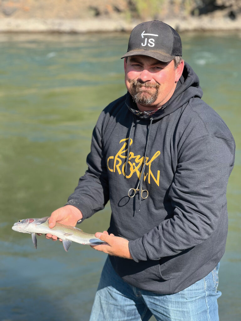 Man in a gray "Royal Crown" hoodie and black JS cap holds a freshly caught trout beside a river