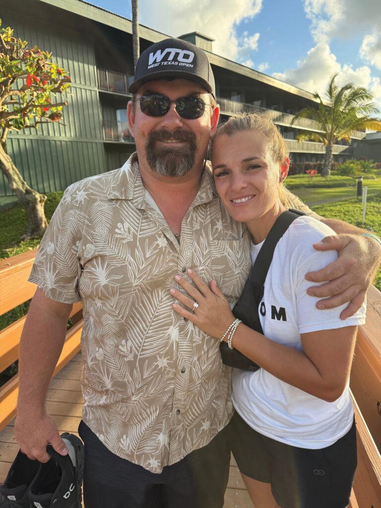 Man in a WTO West Texas Open cap and tan floral shirt hugs a smiling woman in a white tee outside a resort