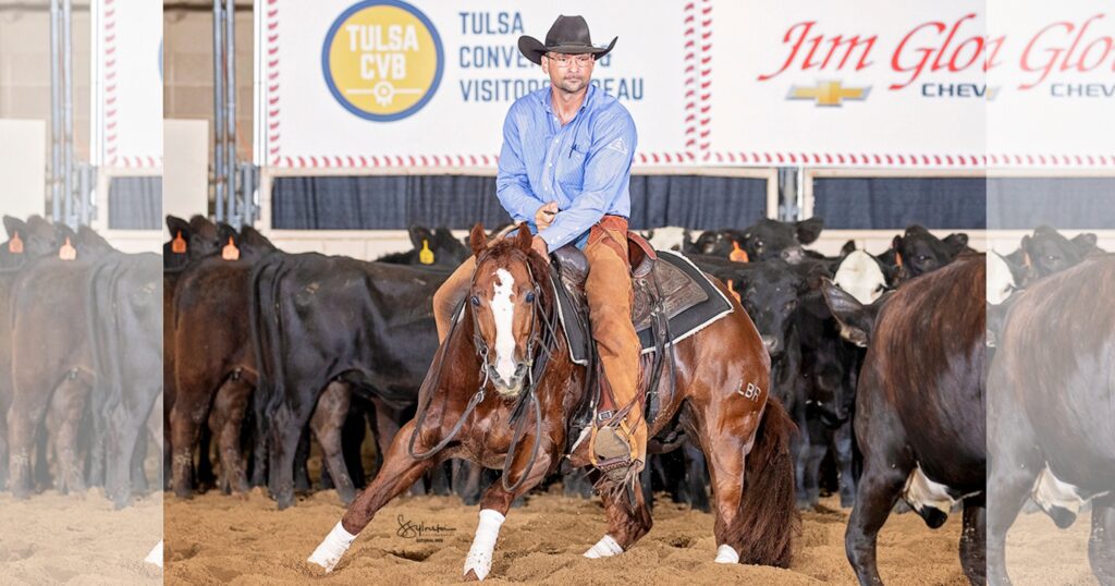 Cutting horse rider in black hat works a sorrel horse low in the dirt in front of a herd of black cattle at an indoor arena