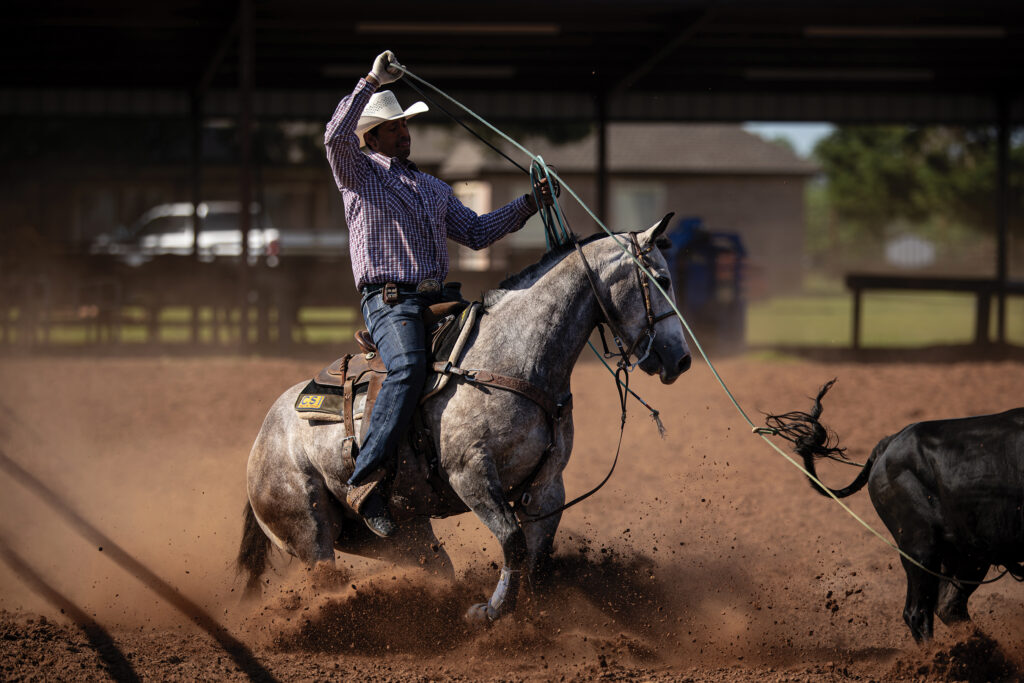 Header roper in a white cowboy hat and plaid shirt pulls his rope tight on a gray horse sliding in the dirt beside a black steer