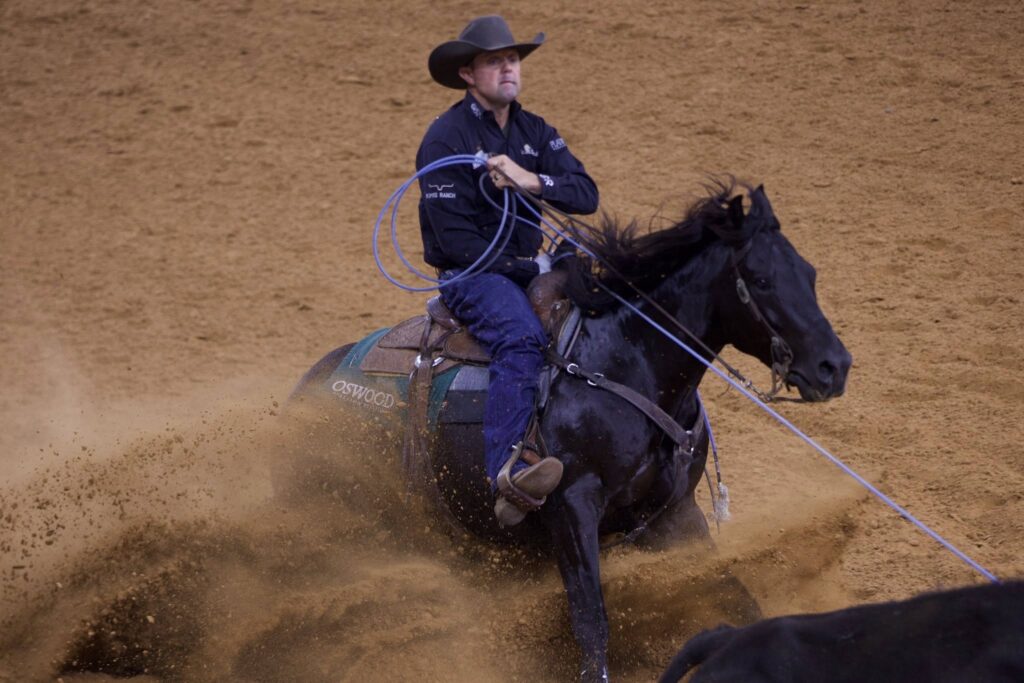 Cowboy in a black hat and navy shirt holds a taut rope as his black horse slides to a stop in the arena dirt