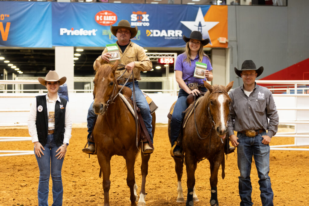 Two mounted riders hold up prize packages in an indoor arena while a woman and man in cowboy hats stand alongside them