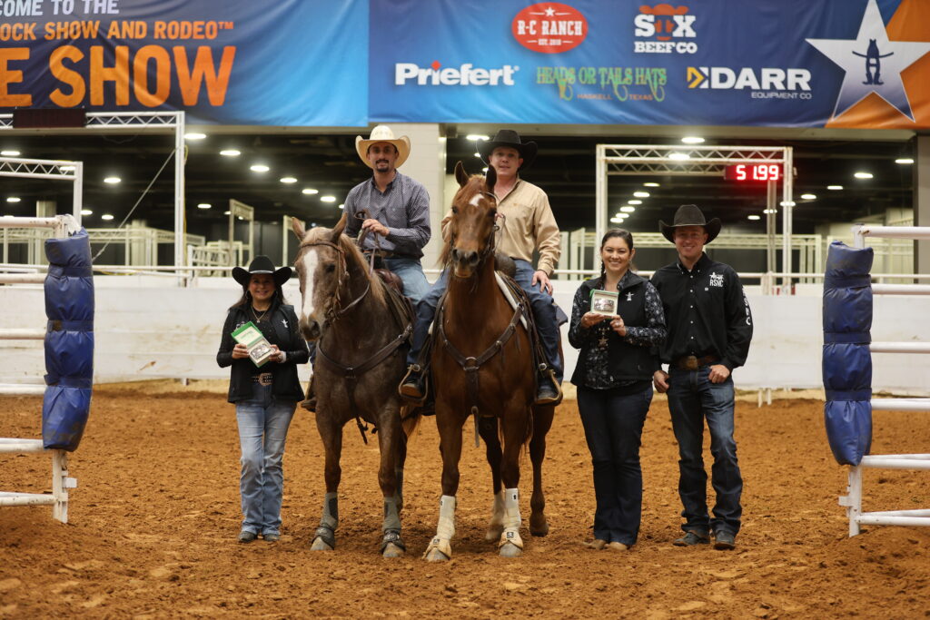 Two mounted riders in cowboy hats pose with two people holding award buckles in an indoor arena beneath rodeo sponsor banners