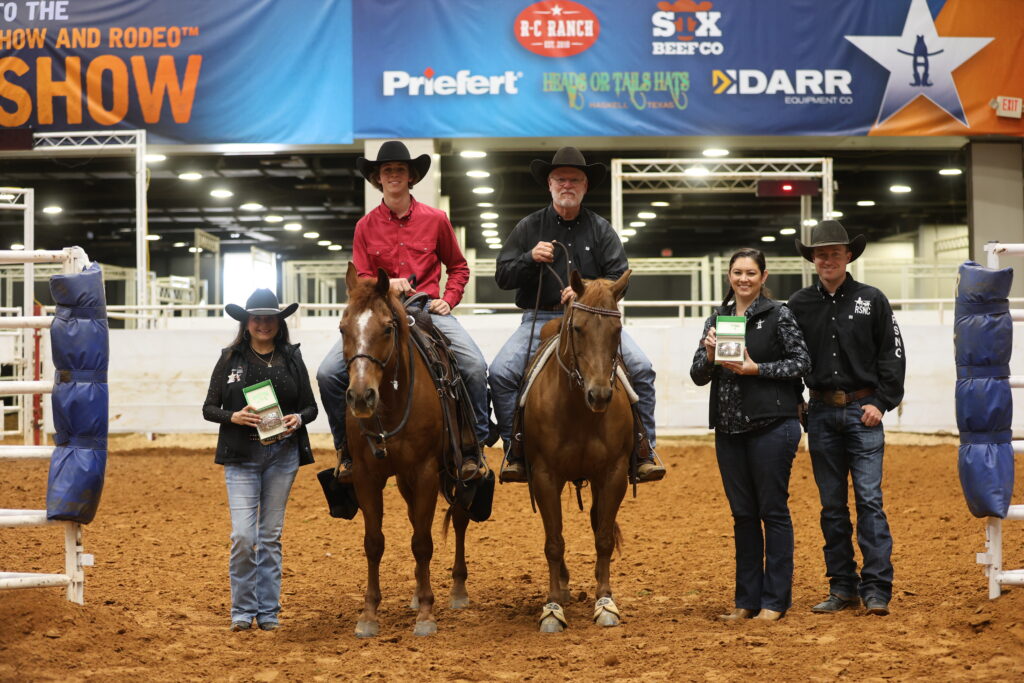 Two mounted riders in cowboy hats pose in an indoor arena with presenters holding award plaques beside them