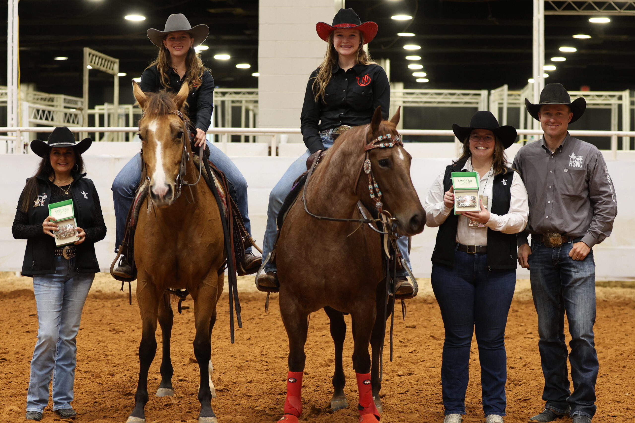 Two young riders in cowboy hats sit horseback in an indoor arena beside three people holding trophy buckles