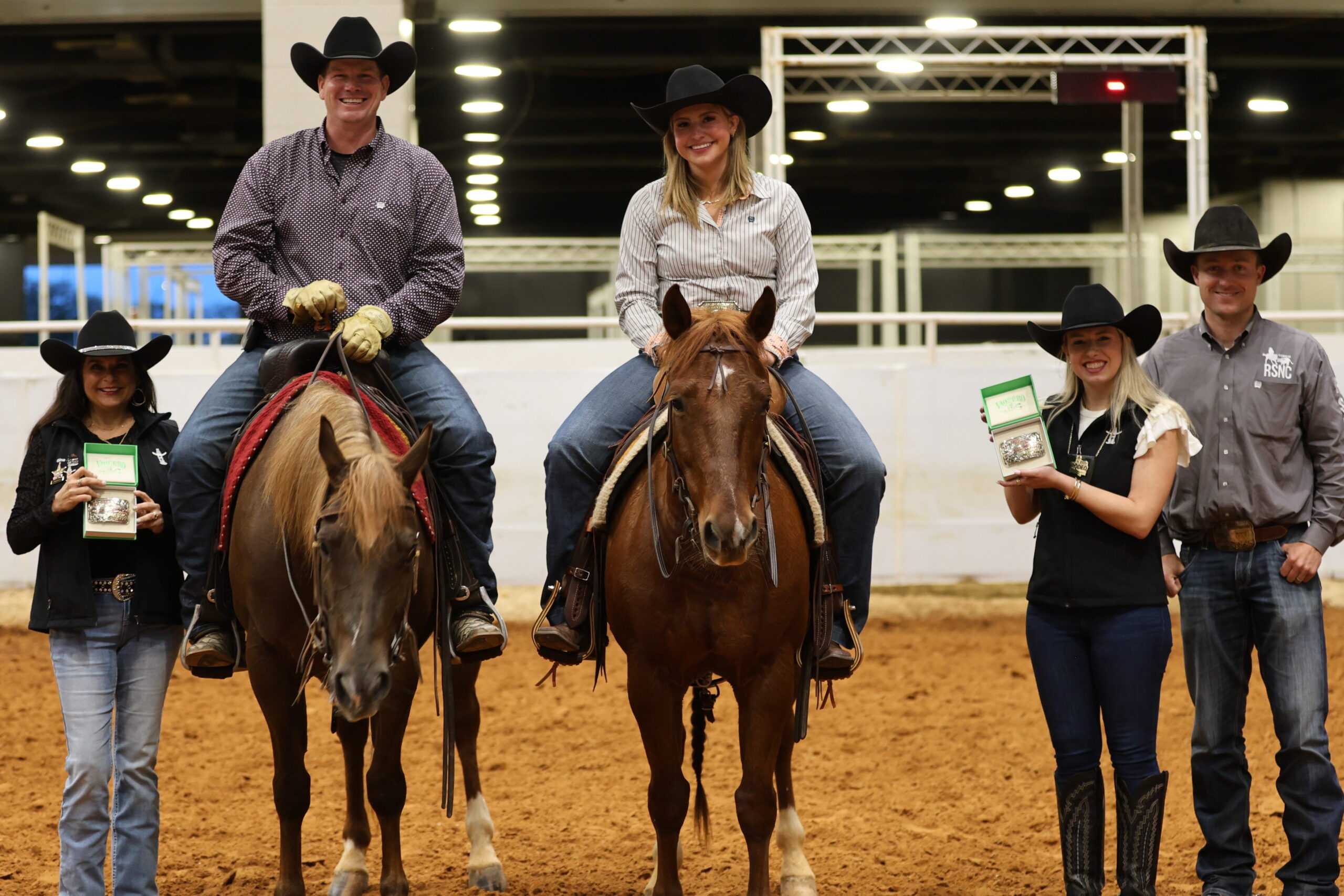 Two mounted riders in black cowboy hats pose in an indoor arena as presenters hold up trophy buckles in green boxes