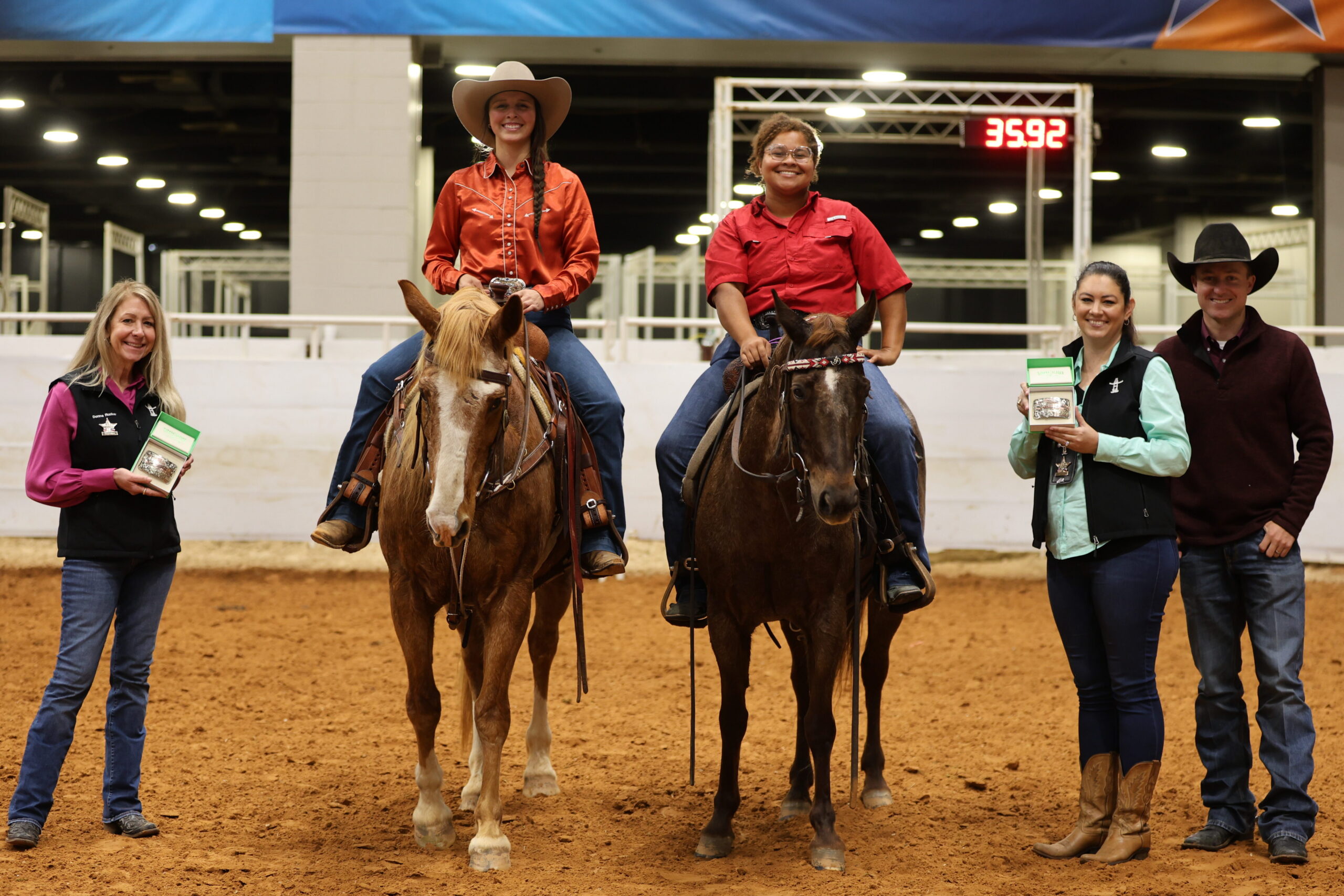 Two mounted riders pose in an indoor arena beside sponsors holding trophy buckles, with 35.92 shown on the timer