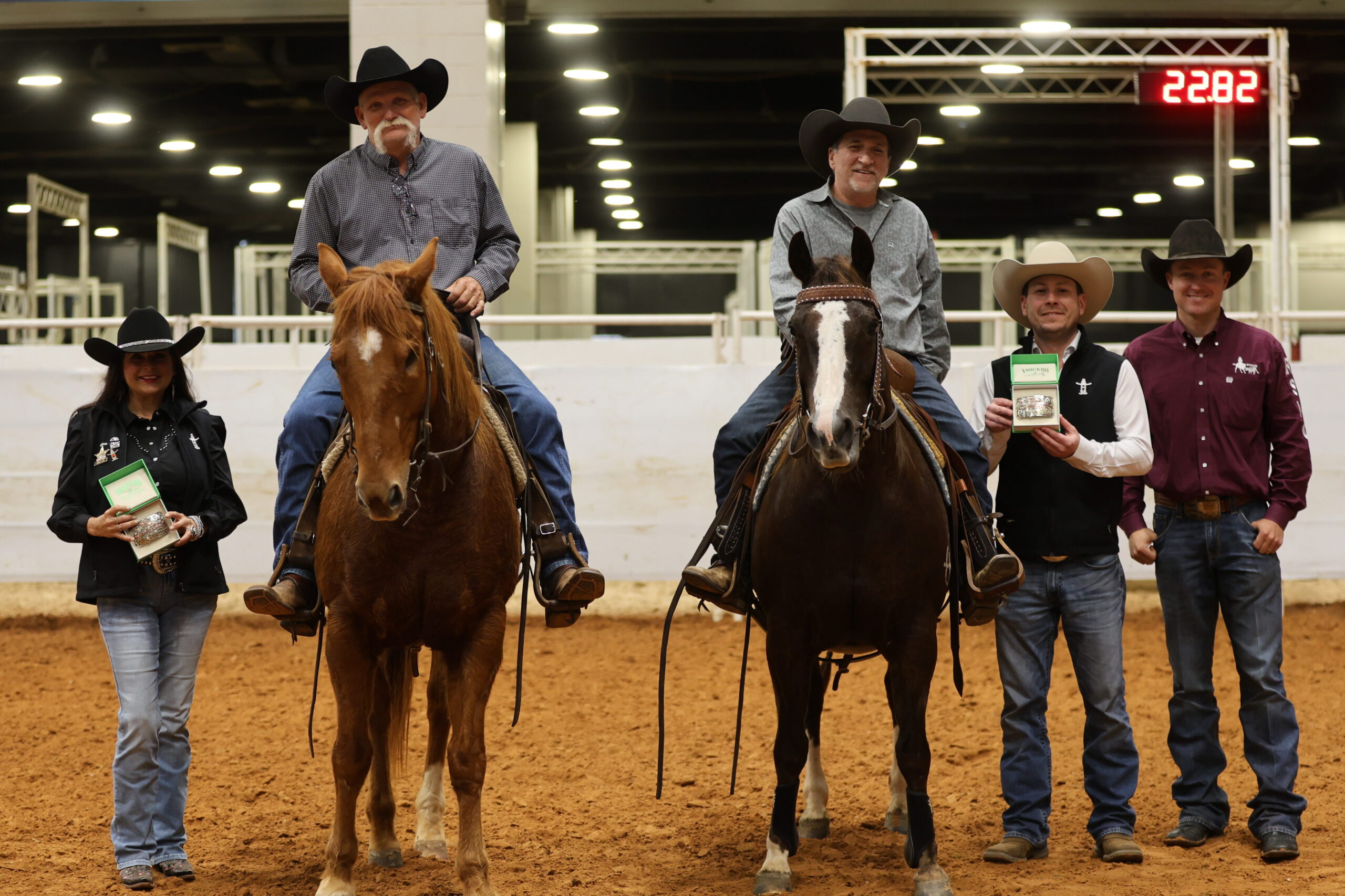 Two mounted team ropers pose in an indoor arena beside presenters holding trophy buckles, with the timer showing 22.82