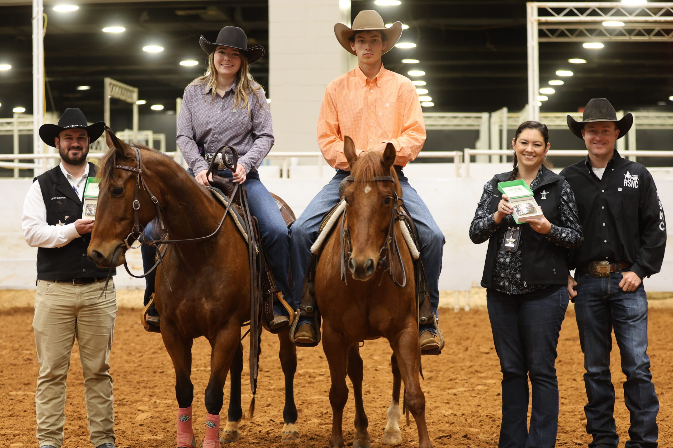 Two young riders on horseback in an indoor arena flanked by RSNC officials presenting championship buckles