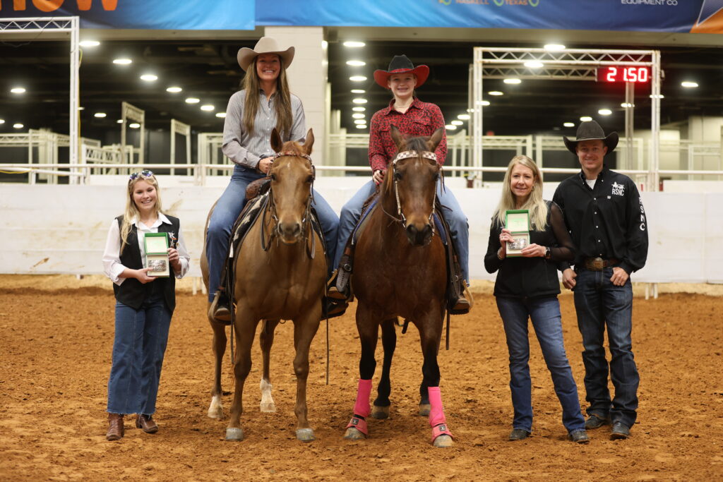 Two riders on horseback in cowboy hats pose in an arena beside three people holding award buckle cases