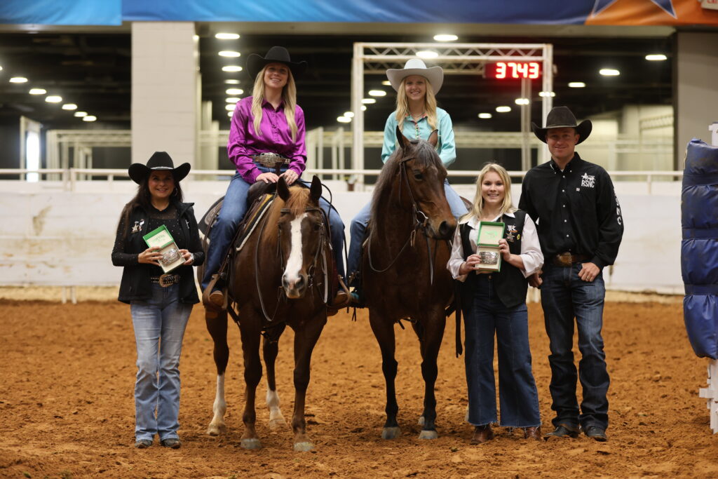 Two riders on horseback pose with three award recipients holding buckle cases in an indoor arena