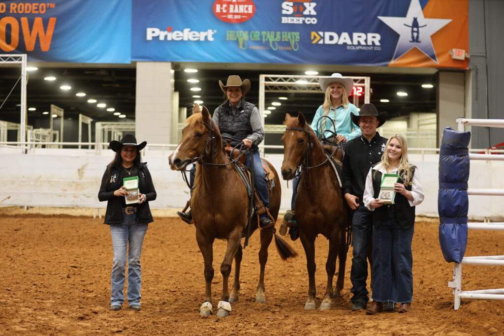 Two riders on horseback pose beside three people holding awards in an indoor arena under rodeo sponsor banners